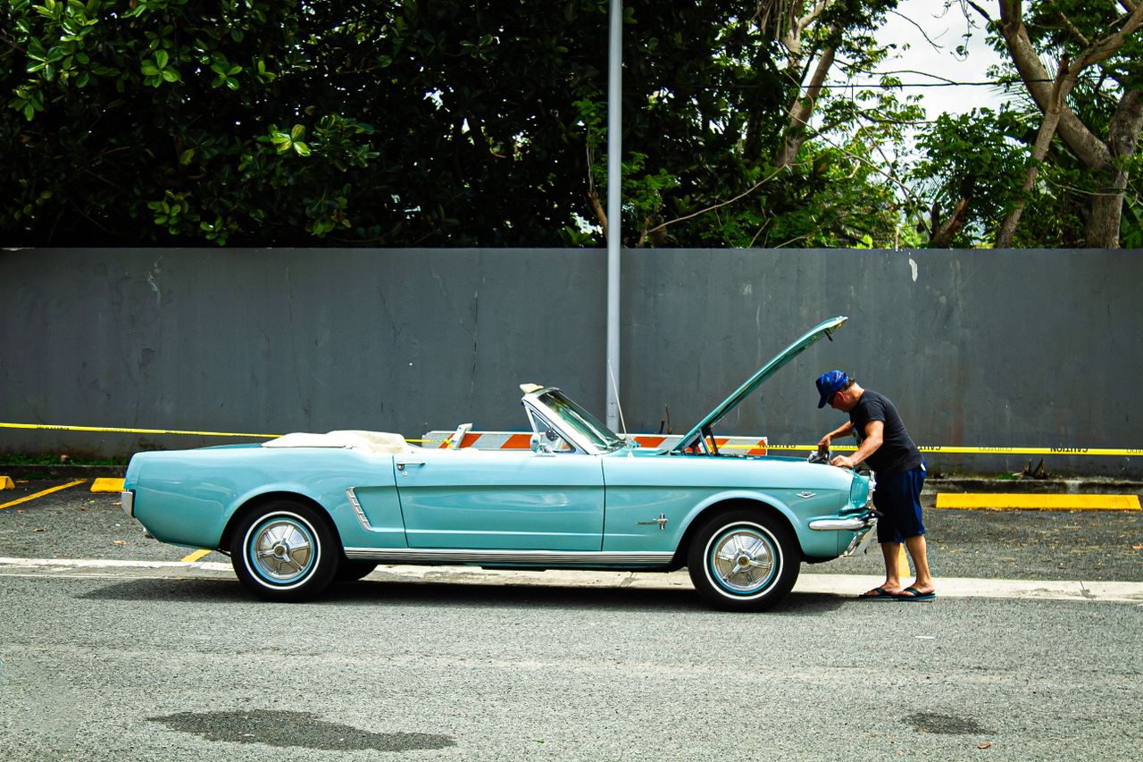 A man checks a vintage blue convertible parked on a street, displaying a classic, nostalgic scene outdoors.